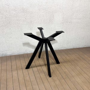 Könnte beinhalten: Ein schwarzes Metall-Tischgestell mit Sternmuster. Das Gestell hat sechs Beine, die sich von einer zentralen Nabe nach außen neigen, mit flachen Montageplatten oben. Das Gestell ist etwa 61 cm hoch.