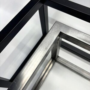 May include: Two metal frames, one black and one silver, are stacked on top of each other. The frames are made of square tubing and are joined at the corners.