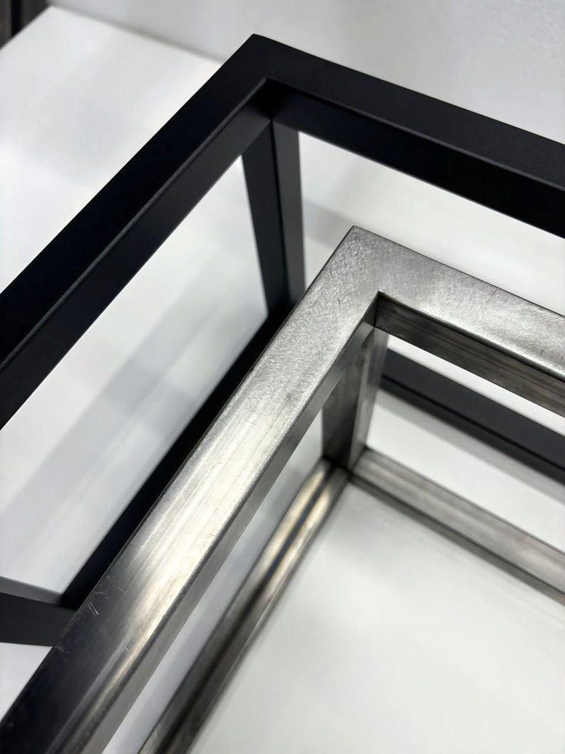May include: Two metal frames, one black and one silver, are stacked on top of each other. The frames are made of square tubing and are joined at the corners.