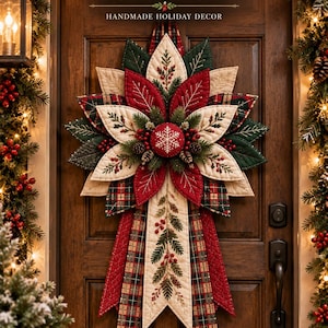 May include: A quilted Christmas door hanger featuring a star-shaped poinsettia design with red, cream, and plaid fabric petals. The centre has a red button with a white snowflake, surrounded by pine sprigs, berries, and long fabric tails.