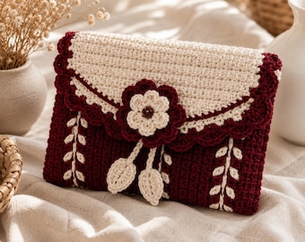 May include: A rectangular crochet pouch in deep burgundy and cream yarn. The flap features a scalloped edge and a central cream flower with burgundy petals. Two cream leaf-shaped charms hang from the centre. The body has vertical cream vine patterns.