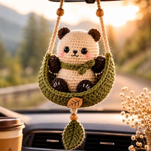 May include: A crocheted panda bear with cream and dark brown yarn, wearing a green scarf, sitting in a green crocheted swing. The swing features a wooden leaf-shaped charm and a dangling green crocheted leaf, suspended by two beige ropes.
