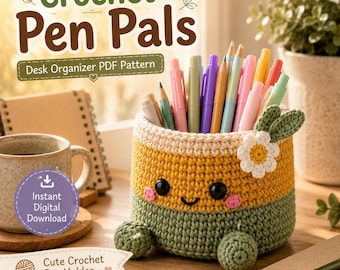May include: A cylindrical crochet pen holder featuring a yellow top section, a green bottom section, and two small green feet. The holder has a smiling face with black eyes and pink cheeks, decorated with a white flower and green leaves.