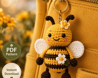 May include: A crocheted bee keyring featuring a yellow and black striped body, white wings, and two small white flowers attached to the head and torso. The bee has black bead eyes, pink cheeks, and is attached to a gold-toned metal keyring.