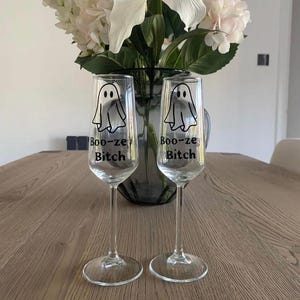 May include: Two clear champagne flutes with a ghost graphic and the text "Boo-ze Bitch". The glasses are on a wooden table, with a vase of white and pink flowers in the background. The flutes are ideal for Halloween.