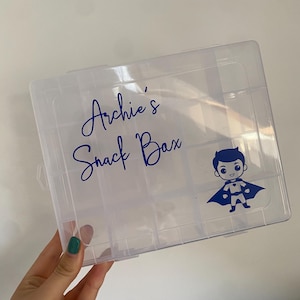 May include: Clear plastic snack box with compartments, labelled "Archie's Snack Box" in blue script, and a superhero graphic. The box is held by a hand with a green painted fingernail.