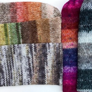 May include: Close-up of several knitted socks in various color combinations. The socks display a range of colors, including orange, brown, green, purple, and gray. The knitted texture is clearly visible, showcasing the craftsmanship of the socks.