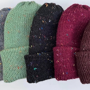 May include: A collection of knitted beanies in various colors, including gray, green, black, burgundy, and brown. Each beanie features a folded cuff and is speckled with colorful flecks. The hats are made of a thick, textured yarn.