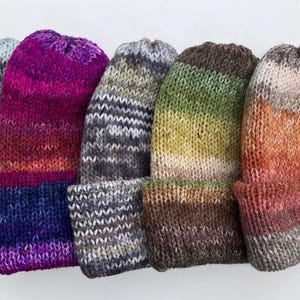 May include: A collection of six knitted beanies in various colors. The beanies feature a folded brim and a gradient color scheme, ranging from dark teal to purple, gray, green, and orange. The knit texture is visible.