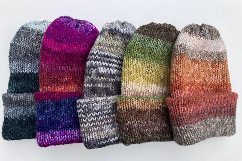 May include: A collection of six knitted beanies in various colors. The beanies feature a folded brim and a gradient color scheme, ranging from dark teal to purple, gray, green, and orange. The knit texture is visible.