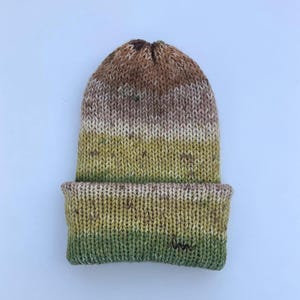 May include: A hand-knitted beanie hat with a folded brim. The hat features a color gradient, transitioning from brown at the crown to green at the brim. The knit pattern is visible, and the hat appears soft and warm.