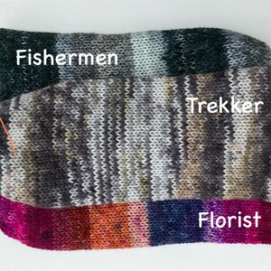 May include: Three pairs of colorful knitted socks are stacked on a white surface. The top pair is labeled "Fishermen" and features green, gray, and white stripes. The middle pair, "Trekker," has a mix of gray, brown, and olive tones. The bottom pair, "Florist," displays pink, orange, and blue hues.