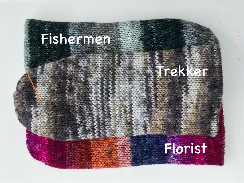 May include: Three pairs of colorful knitted socks are stacked on a white surface. The top pair is labeled "Fishermen" and features green, gray, and white stripes. The middle pair, "Trekker," has a mix of gray, brown, and olive tones. The bottom pair, "Florist," displays pink, orange, and blue hues.