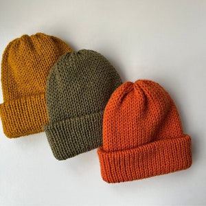 Puede incluir: Tres gorros de punto en tonos amarillo mostaza, verde oliva y naranja. Los sombreros están hechos de un hilo grueso y texturizado con un borde doblado. Los gorros están dispuestos en diagonal, mostrando sus colores y texturas. Ideal para otoño e invierno.
