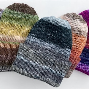 May include: A collection of five knitted beanies in various colors. The hats feature a folded brim and a gradient color scheme, with hues ranging from gray and green to orange and purple. The beanies are made of a soft, textured yarn.