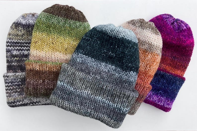 May include: A collection of five knitted beanies in various colors. The hats feature a folded brim and a gradient color scheme, with hues ranging from gray and green to orange and purple. The beanies are made of a soft, textured yarn.