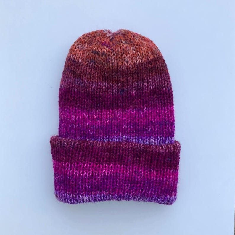May include: A hand-knitted beanie hat in a gradient of colors, transitioning from orange at the crown to shades of red and purple at the brim. The hat is made of a soft, textured yarn.