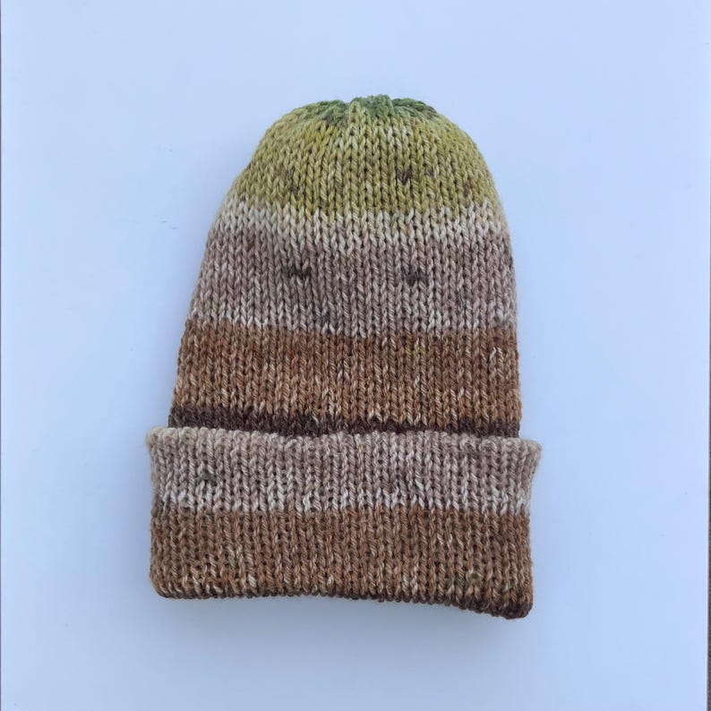 May include: A hand-knitted beanie hat in shades of brown, tan, and green. The hat features a folded brim and a textured knit pattern. The color gradient transitions from green at the crown to brown at the brim.