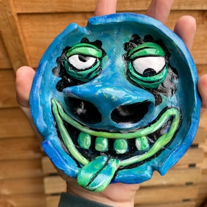 May include: A hand-painted, blue ceramic ashtray with a quirky face design. It has green eyes, a green tongue, and a wide, grinning mouth. The ashtray is circular with a slightly raised edge.