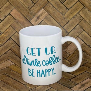 Könnte beinhalten: Eine weiße Keramik-Kaffeetasse mit einer türkisfarbenen Aufschrift, die "Get Up. Drink Coffee. Be Happy." lautet.