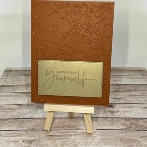 Könnte beinhalten: Eine braune Karte mit einem Blumenmuster und einem goldenen Rechteck mit dem Text "Always be yourself" darauf. Die Karte wird auf einem Holzständer präsentiert.