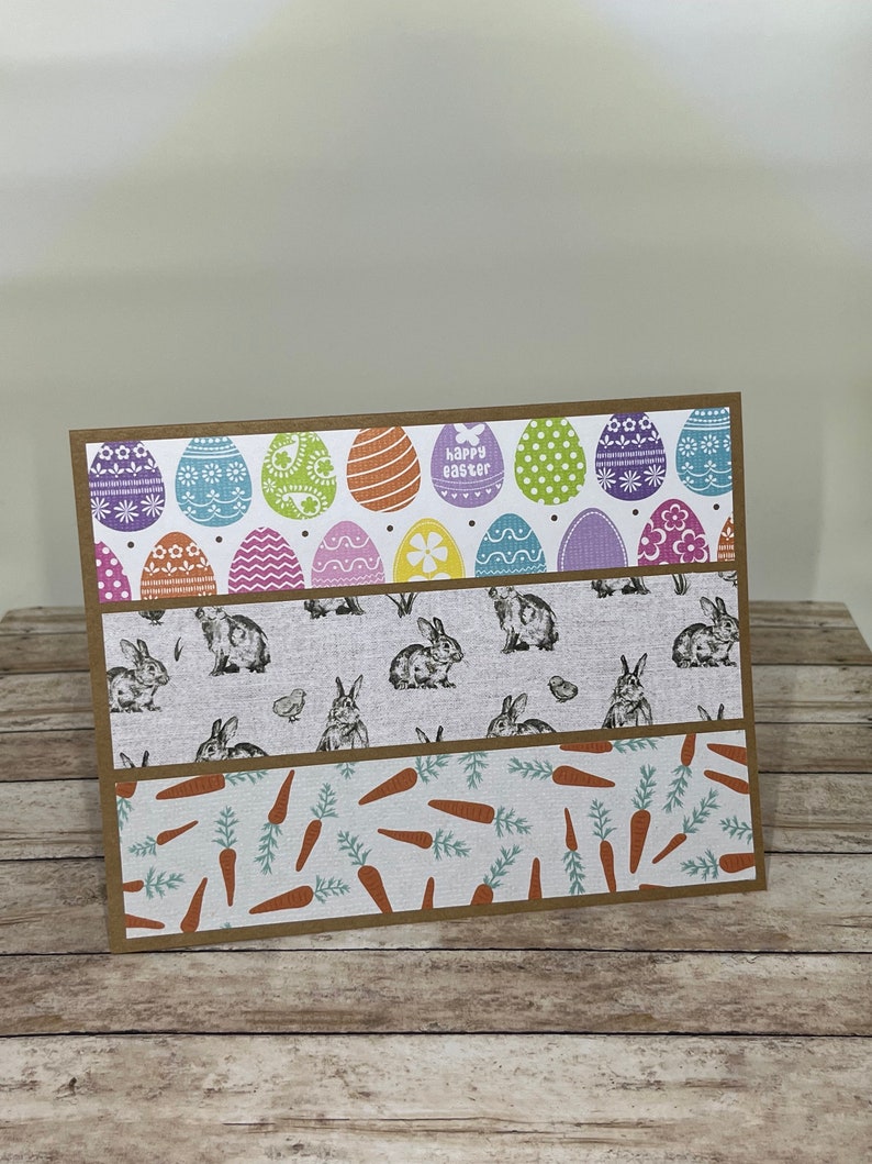 May include: Easter greeting card with three patterned sections. The top section features colorful Easter eggs with the text "happy easter". The middle section has rabbit illustrations, and the bottom section has carrot illustrations. The card has a brown border.