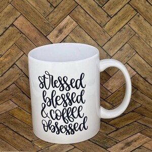 Könnte beinhalten: Weiße Keramik-Kaffeetasse mit schwarzem Text, der "gestresst gesegnet & kaffeebesessen" lautet.