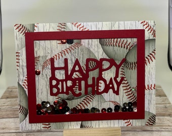May include: A handmade birthday card with a baseball theme. The card features a red frame with the words "HAPPY BIRTHDAY" cut out. The background has a baseball pattern and decorative sequins.