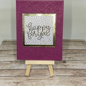 Könnte beinhalten: Eine rosa Karte mit goldenem Rahmen und weißen Punkten. Die Karte sagt "happy for you" in goldenen Buchstaben. Die Karte steht auf einem kleinen Holzständer.