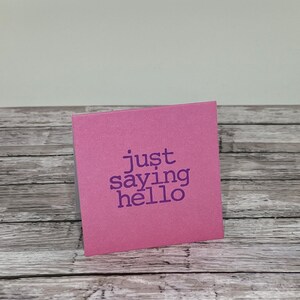 May include: A square, pink greeting card with the phrase "just saying hello" printed in purple. The card is set against a backdrop of a wooden surface and a plain white wall.