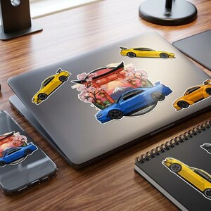 May include: A laptop, phone, and notebook are decorated with car stickers. The stickers depict sports cars in yellow, blue, and orange, alongside a Japanese-style graphic. A keyboard, mouse, and coffee mug are also present.