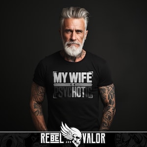 Puede incluir: Camiseta negra con el texto "MY WIFE IS PSYCHOTIC" en un diseño blanco y gris. La imagen presenta a un hombre con pelo y barba grises, que lleva la camiseta. El logotipo de Rebel and Valor está en la parte inferior.