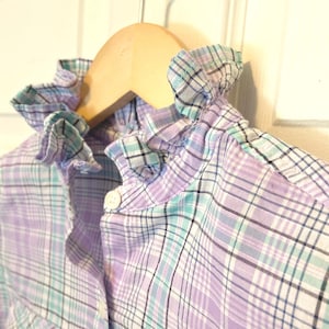 May include: A light purple plaid shirt with a ruffled collar, hanging on a wooden hanger. The shirt features a button-down front and a pattern of purple, blue, and black lines. The collar has a ruffled edge.