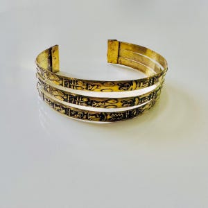 Puede incluir: Brazalete de puño dorado con tres bandas. Cada banda presenta una serie de símbolos jeroglíficos oscuros grabados. El brazalete tiene una apariencia ligeramente envejecida, lo que sugiere un estilo antiguo o vintage. El diseño evoca la estética del antiguo Egipto.