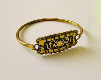 May include: A gold-toned bangle bracelet with a rectangular, ornate centerpiece. The centerpiece features a black background with gold-colored, stylized designs. The bracelet has a simple, circular band and a clasp closure.