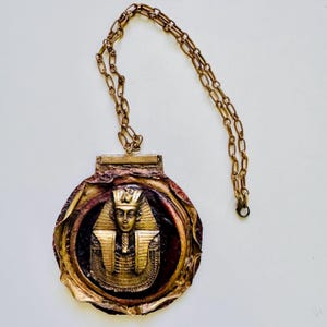 Puede incluir: Un collar con colgante vintage con un diseño de faraón dorado. El colgante circular presenta un relieve detallado del rostro y el tocado de un faraón. El collar tiene una larga cadena de eslabones.