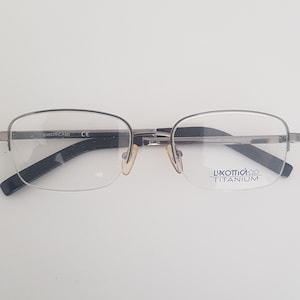 May include: Silver-toned spectacles with black arms and clear lenses. The right lens has the text "Luxottica Titanium" printed on it.