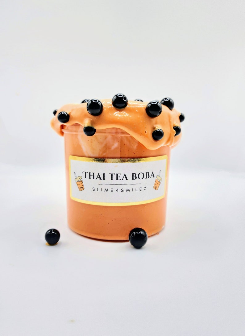 K&ouml;nnte beinhalten: Orangener Schleim mit schwarzen Boba-Perlen in einem durchsichtigen Glas mit einem goldenen Etikett, auf dem "THAI TEA BOBA SLIME4SMILEZ" steht.