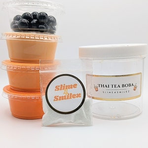 K&ouml;nnte beinhalten: Ein durchsichtiger Plastikbeh&auml;lter gef&uuml;llt mit schwarzen Boba-Kugeln, ein kleiner Beutel mit wei&szlig;em Schleimpulver mit einem Etikett, auf dem "Slime 4 Smilez" steht, und ein durchsichtiges Plastikglas mit wei&szlig;em Deckel und einem Etikett, auf dem "Thai Tea Boba Slime 4 Smilez" steht.