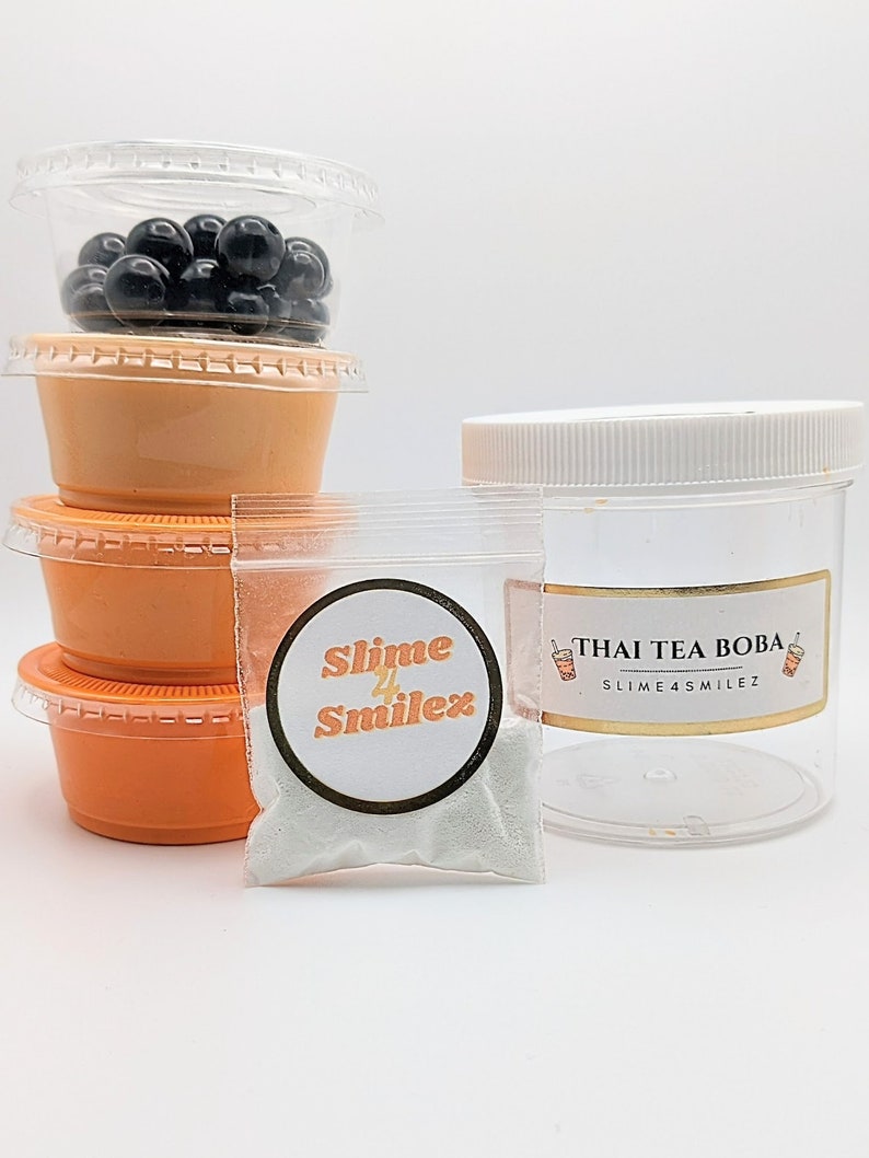 K&ouml;nnte beinhalten: Ein durchsichtiger Plastikbeh&auml;lter gef&uuml;llt mit schwarzen Boba-Kugeln, ein kleiner Beutel mit wei&szlig;em Schleimpulver mit einem Etikett, auf dem "Slime 4 Smilez" steht, und ein durchsichtiges Plastikglas mit wei&szlig;em Deckel und einem Etikett, auf dem "Thai Tea Boba Slime 4 Smilez" steht.