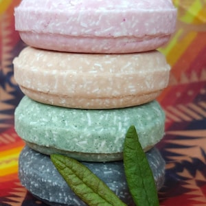 Pode incluir: Uma pilha de quatro barras de champô sólidas redondas, em tons de rosa, pêssego, verde e cinza. As barras têm um aspeto texturizado, com flocos visíveis. Duas folhas verdes repousam ao lado da barra inferior, sobre um fundo colorido com padrões.