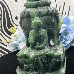 May include: Green jade Buddha statue with intricate details. The Buddha is seated in a meditative pose with hands clasped in front. The statue is carved from a single piece of jade and has a smooth, polished finish.