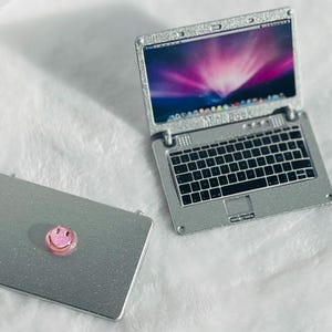 May include: A silver laptop computer with a black keyboard and a smiley face sticker on the lid. The laptop is closed and sitting on a white surface.