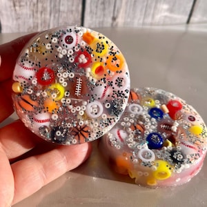 Puede incluir: Dos posavasos redondos de resina con temática deportiva. Los posavasos son transparentes y están llenos de purpurina de colores, cuentas y miniaturas de equipos deportivos, incluyendo pelotas de béisbol, baloncesto, fútbol americano y fútbol.
