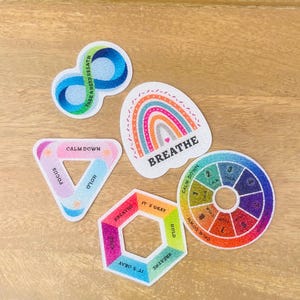 Pode incluir: Cinco autocolantes coloridos com uma textura brilhante. Um é um laço infinito azul, um é um arco-íris com o texto BREATHE, um é um triângulo com texto, um é um hexágono com texto e um é uma roda circular com os números de um a oito.