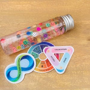 Puede incluir: Un conjunto de herramientas de atención plena de colores que incluye un tubo de plástico transparente lleno de caritas sonrientes de colores, una rueda de colores circular con las palabras "Calm Down" y "Focus" impresas en ella, un triángulo con las palabras "Calm Down", "Focus" y "Hold" impresas en él, y un símbolo de infinito con las palabras "Take a Deep Breath" impresas en él.