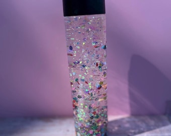 Könnte beinhalten: Eine durchsichtige, zylindrische Sensorik-Flasche mit schwarzem Schraubverschluss, gefüllt mit einer Flüssigkeit, die eine Vielzahl von buntem, unterschiedlich geformtem Glitzer, kleinen Kunststoffperlen und winzigen dekorativen Anhängern enthält.