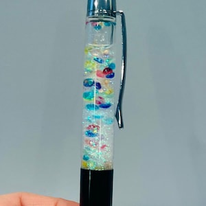 Op de afbeelding: Een zilveren en zwarte pen met een transparant lichaam gevuld met kleurrijke glitter en iriserende kralen. De pen heeft een zilveren clip en een zwarte dop.