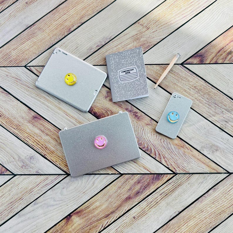 May include: A flat lay of school supplies on a woodgrain surface. The items include a composition book, a pencil, a silver laptop with a pink smiley face sticker, a silver tablet with a yellow smiley face sticker, and a silver phone with a blue smiley face sticker.