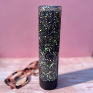 May include: A clear glass cylinder filled with black glitter and iridescent green and silver stars. The cylinder has a black lid and is sitting on a light wood surface.